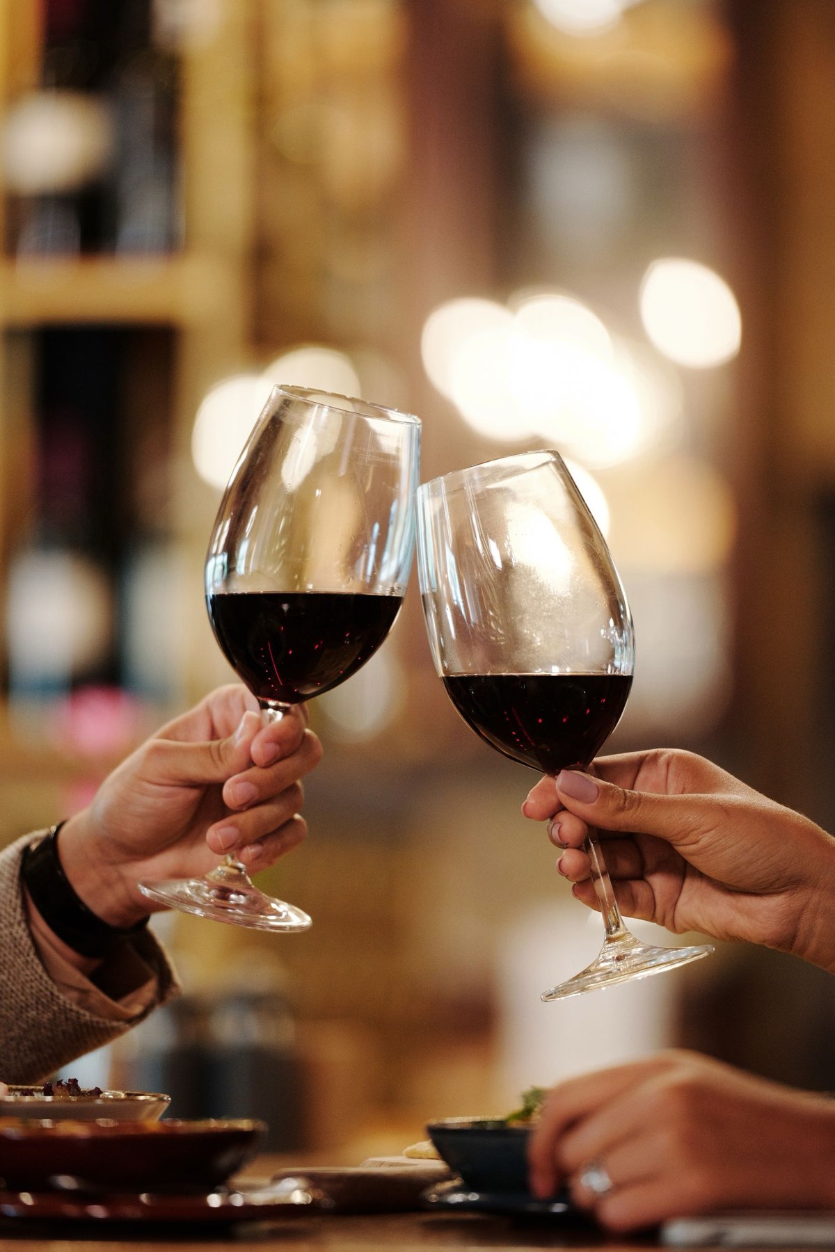Twee handen die met een glas rode wijn proosten aan een tafel bij zacht avondlicht